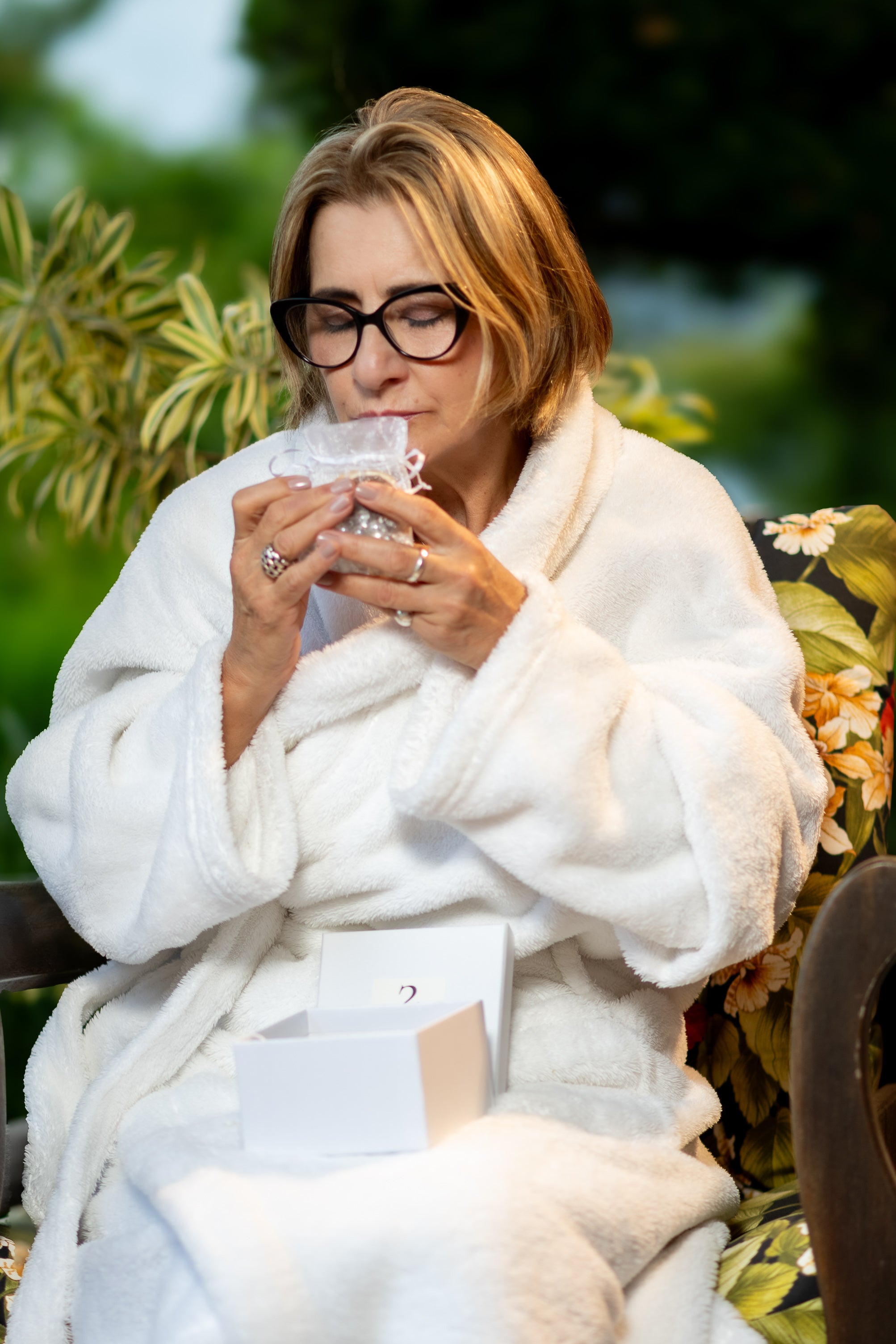 A woman in a white robe holding a small fragrance sachet to her face, a JU4U box on her lap