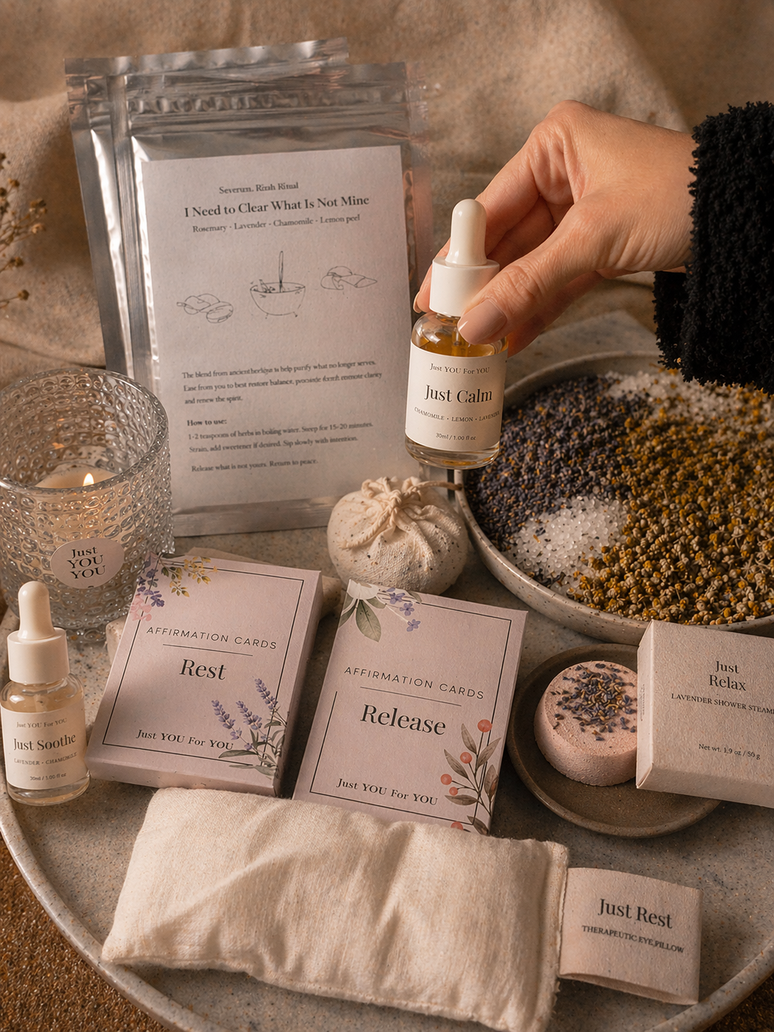 A hand holding a Just Calm oil over a bowl of herbal salts, surrounded by affirmation cards, a candle and an eye pillow